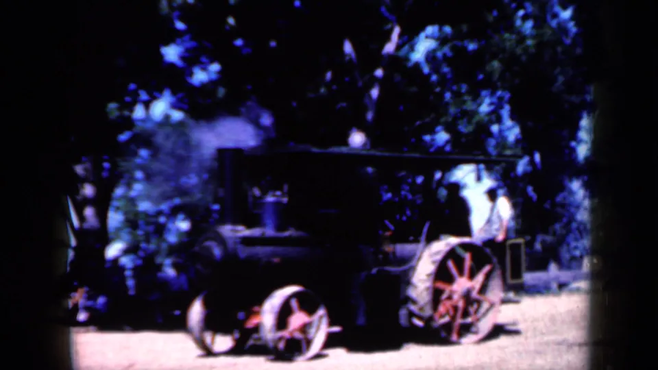 1959 Iowa 8mm frame showing a dark steam tractor with red-spoked wheels outdoors under trees as pale steam rises beside it.
