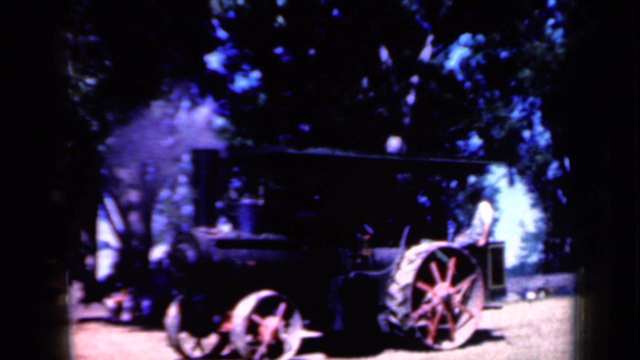 1959 Iowa home-movie frame of a black steam tractor with red wheels, pale steam, trees, and a person visible near the machinery.