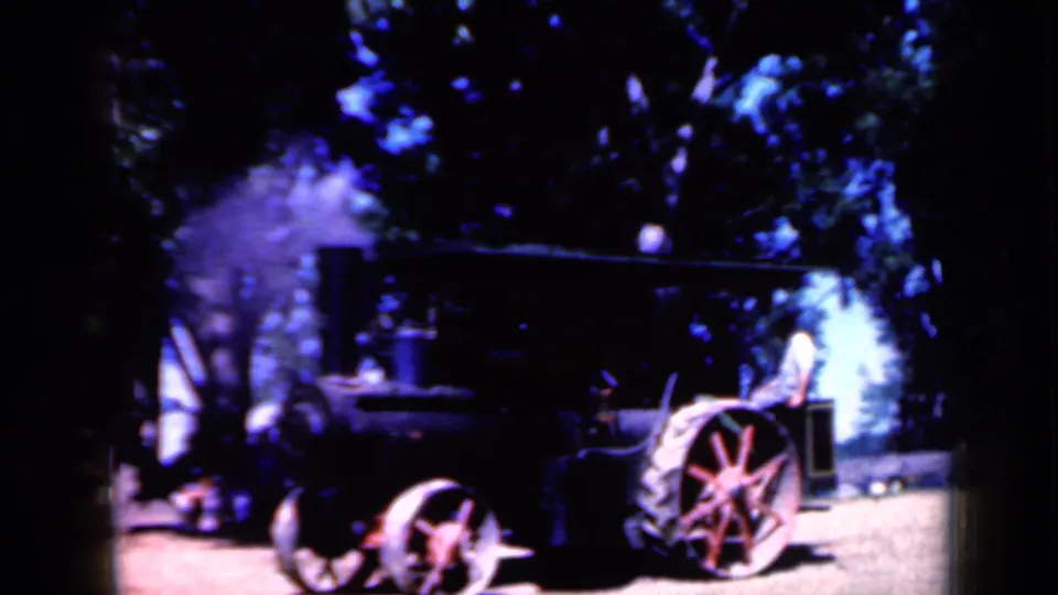 1959 Iowa home-movie frame of a black steam tractor with red wheels, pale steam, trees, and a person visible near the machinery.