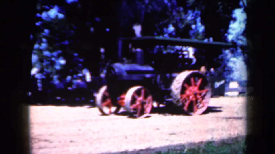 1959 Iowa 8mm frame showing a side view of a dark steam traction engine with red-spoked wheels on a sunlit dirt surface.