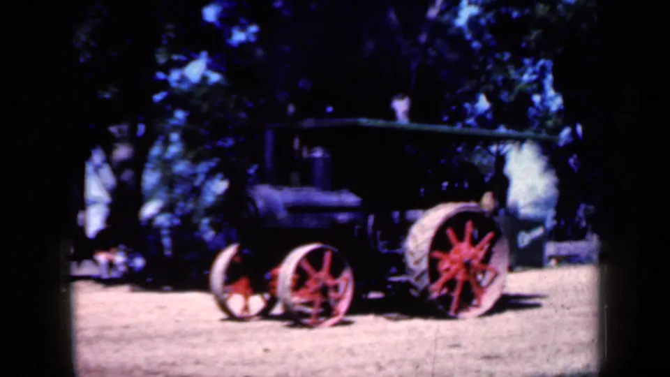 1959 Iowa home-movie frame of the steam tractor in partial side view, with red wheel spokes, dark bodywork, trees, and dirt foreground.