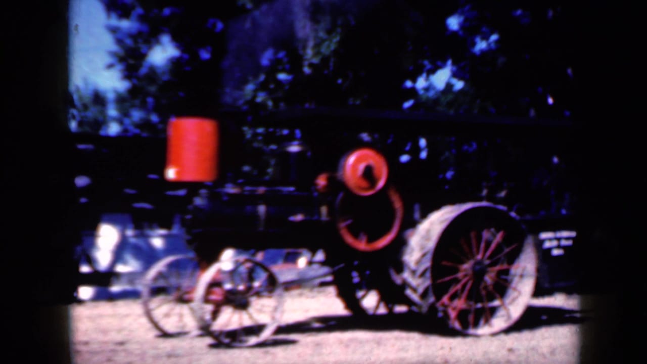 1959 Iowa home-movie frame of a dark steam-tractor style machine with red wheel details, a tall red cylinder, and pale dirt ground beneath trees.