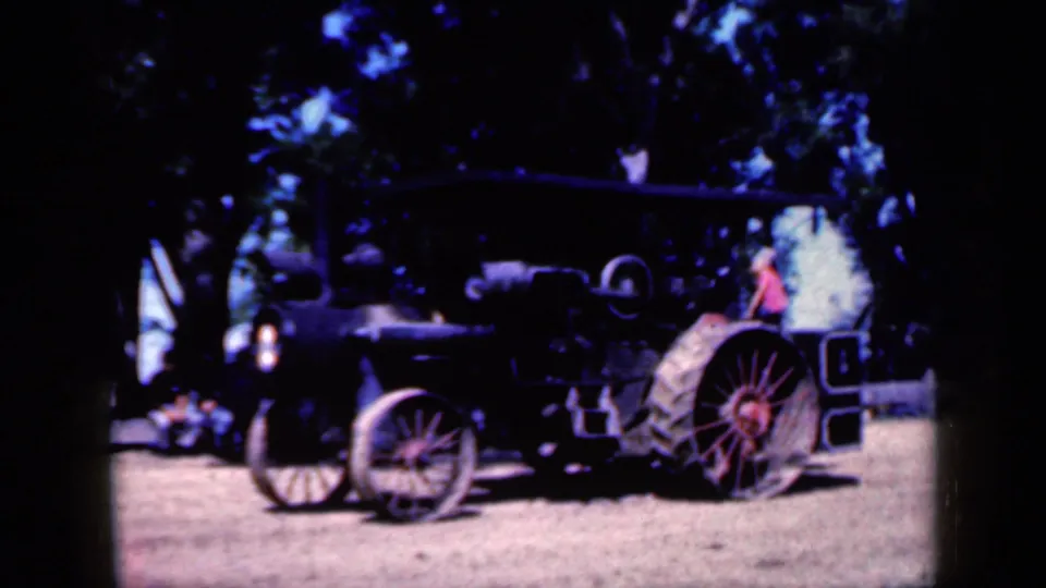 1959 Iowa 8mm frame with a dark antique tractor, red spoked rear wheel, skinny front wheel, and a person in red standing near the rear area.