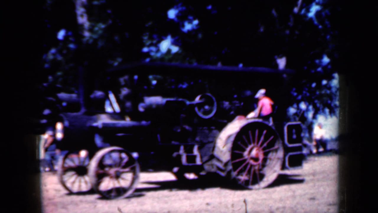Side view of the steam tractor in Iowa, 1959, with a person in a pink shirt and light hat seated near the rear wheel.