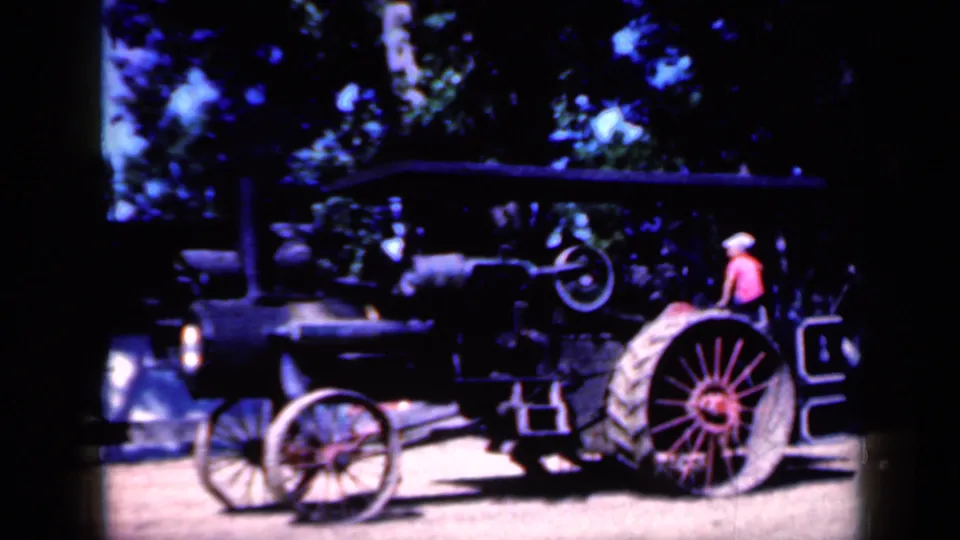 1959 Iowa home-movie frame of a black steam tractor, its canopy, front wheel, large rear wheel, and seated person partly visible.