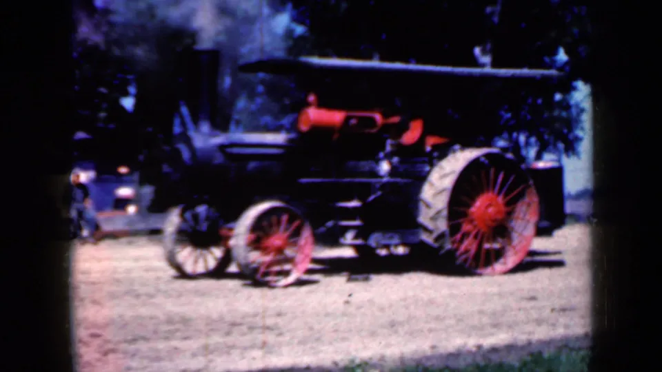 Wider 1959 Iowa frame showing the steam tractor with red-spoked wheels, black body, canopy, smokestack, trees, and a person nearby.