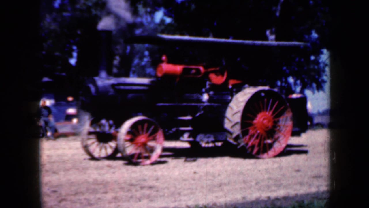 Steam traction engine with red wheels on a dirt field, with trees and distant people behind it.