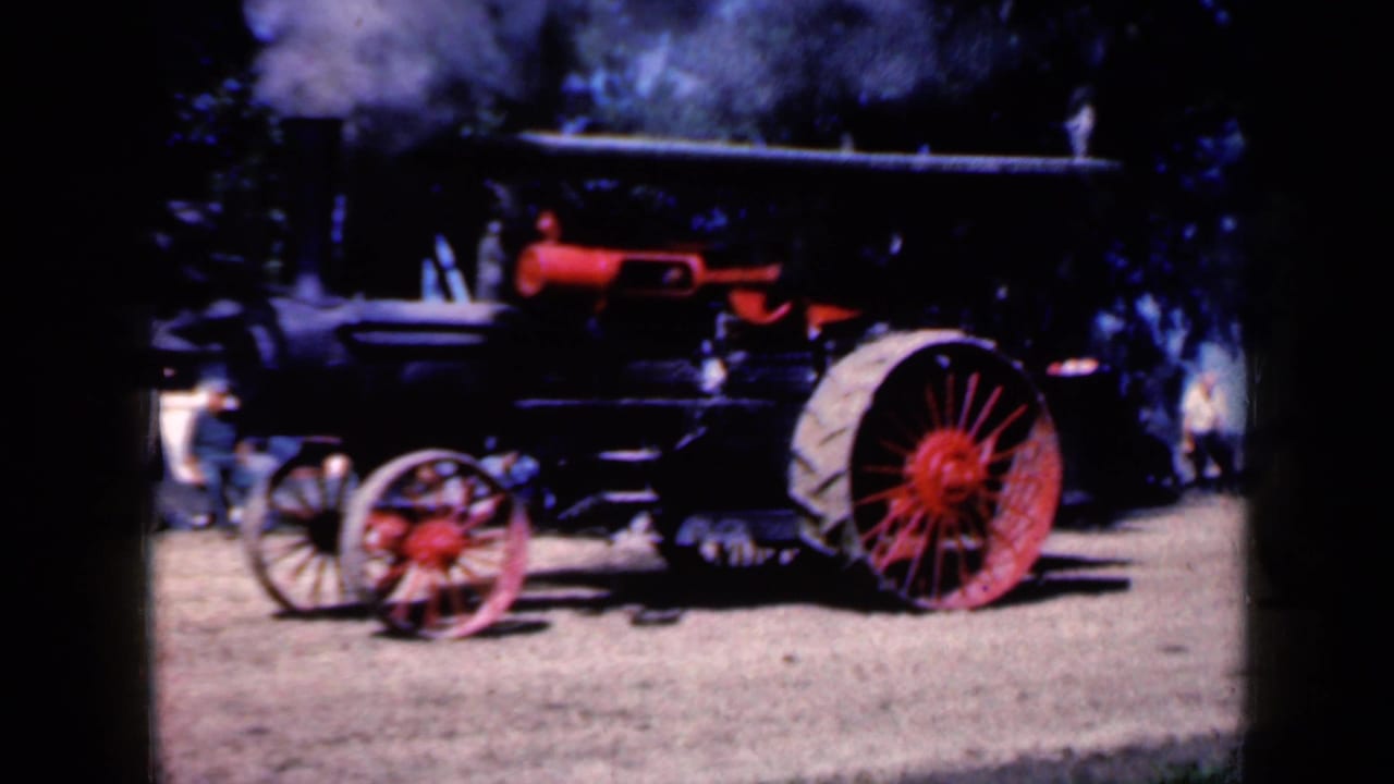1959 Iowa home-movie frame of the steam traction engine in profile, with red wheels, pale rear tire and smoke drifting upward.