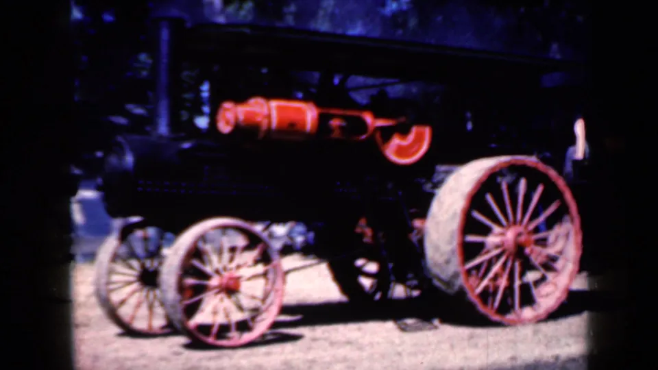 Close 1959 Iowa 8mm view of the traction engine, emphasizing the red wheel spokes, black machinery and sunlit dirt surface.