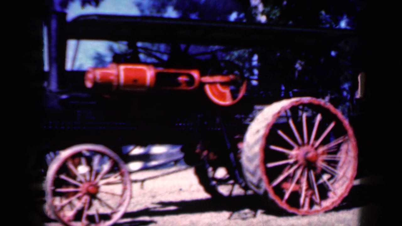 1959 Iowa frame of the steam-powered tractor from a slightly shifted side angle, with red wheel spokes and dark tree cover behind it.