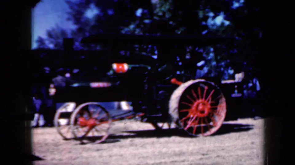 1959 Iowa home-movie frame showing the steam tractor from a wider angle, with red spoked wheel, dark body, trees, and indistinct people nearby.