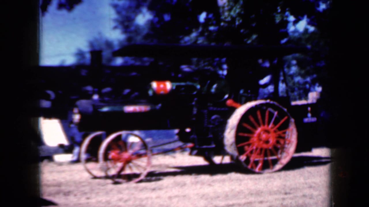 Black steam tractor with red spoked wheels moving across a dirt fairground.