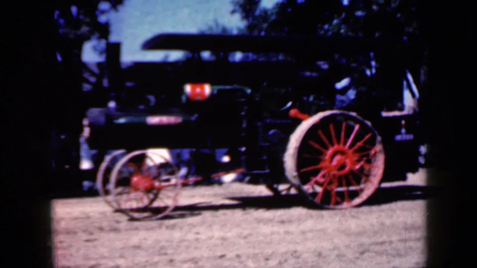 Dark steam tractor with a large red wheel passes through the 1959 Iowa fairground scene on grainy 8mm film.