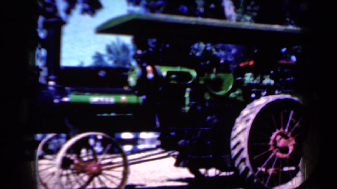 Green steam tractor with large red spoked wheel under trees.