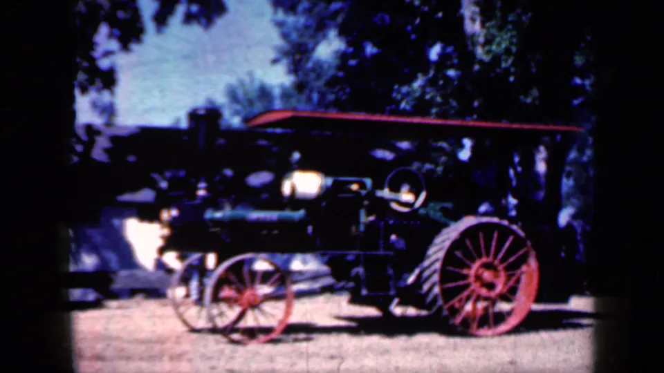 Wider 1959 Iowa 8mm frame showing the green steam tractor in profile on a dirt field with red spoked wheels and trees in the background.