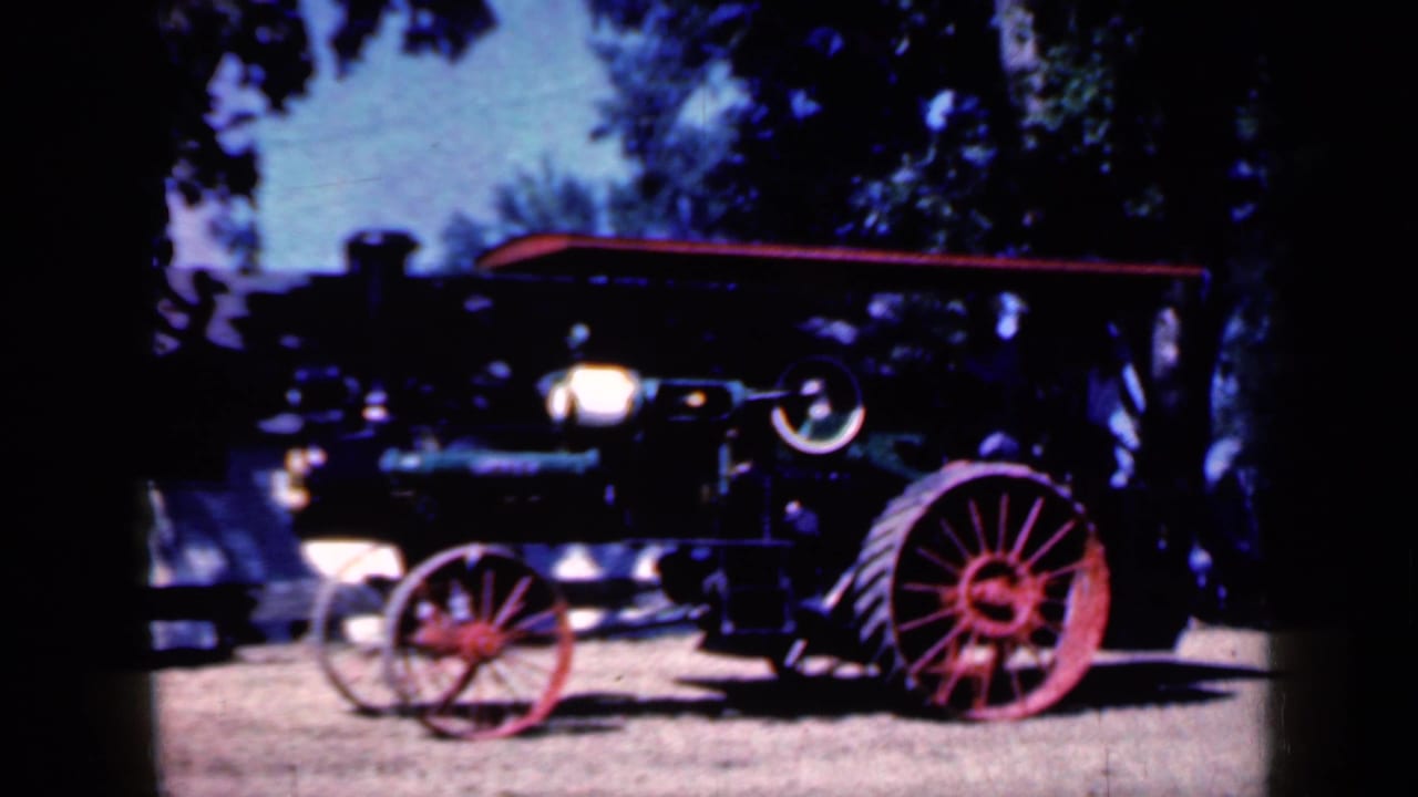 Steam traction engine with red spoked wheels parked on dirt beneath trees.