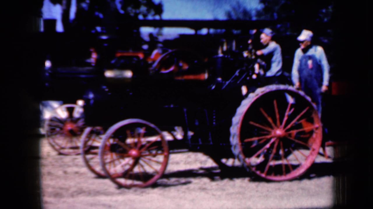 1959 Iowa 8mm frame showing two men near a black steam-powered tractor with red spoked wheels in bright daylight.
