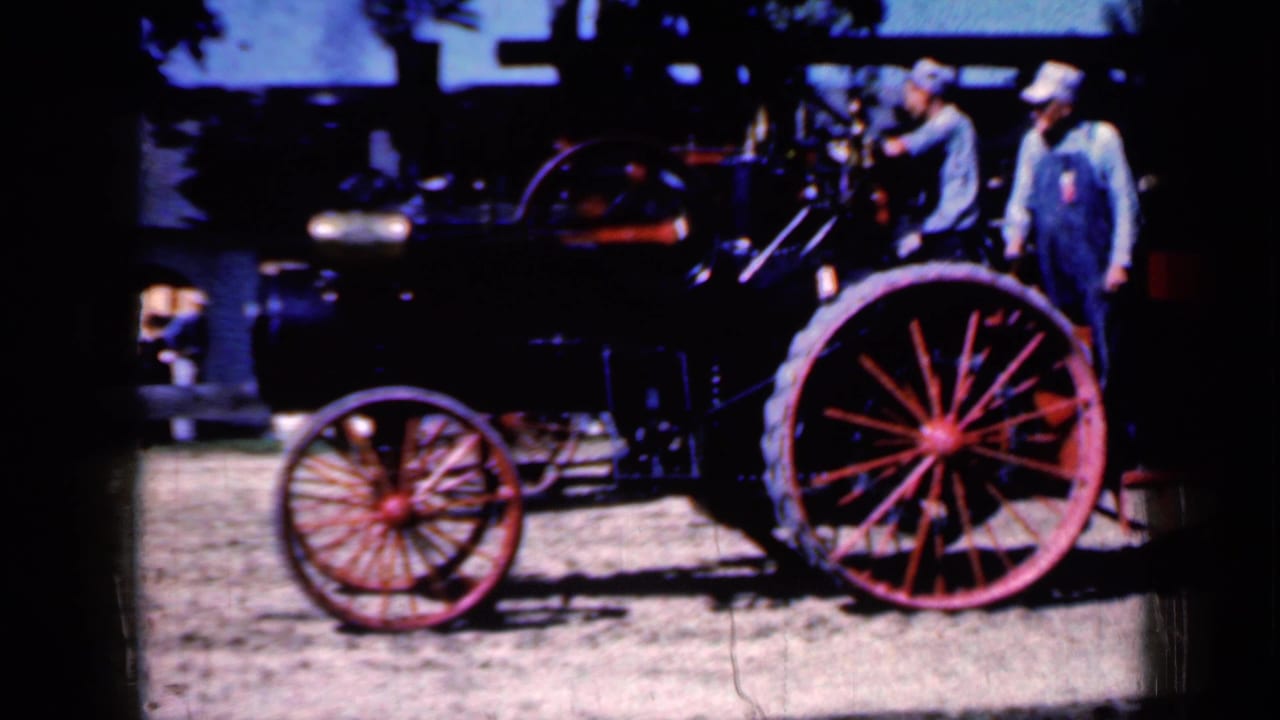 A steam tractor with red spoked wheels is seen from the side with two men nearby.