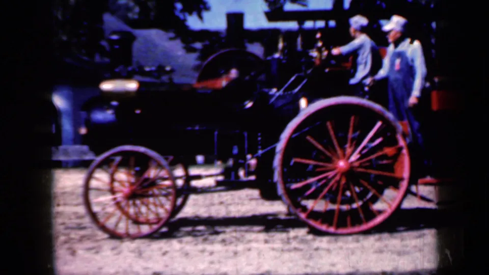1959 Iowa home-movie frame with the black tractor body and red spoked wheels dominating the foreground.