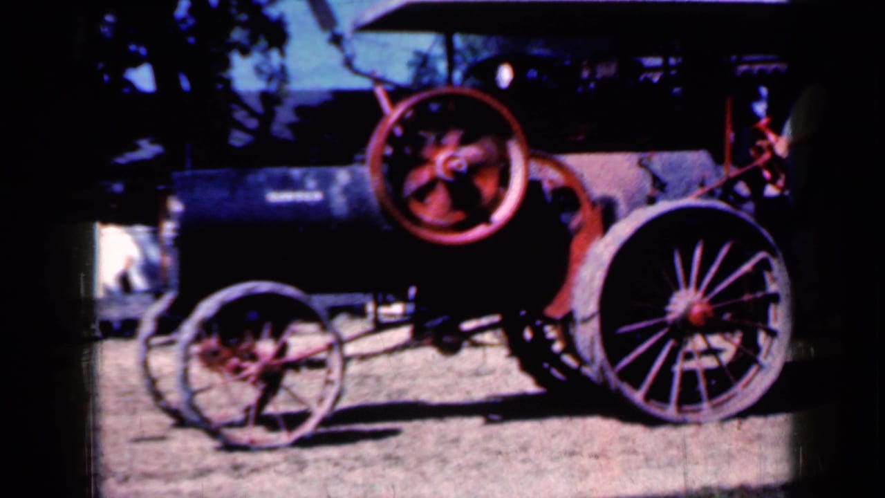 1959 Iowa home-movie frame of a weathered steam tractor, with the red flywheel prominent and a person partly visible near the machinery.