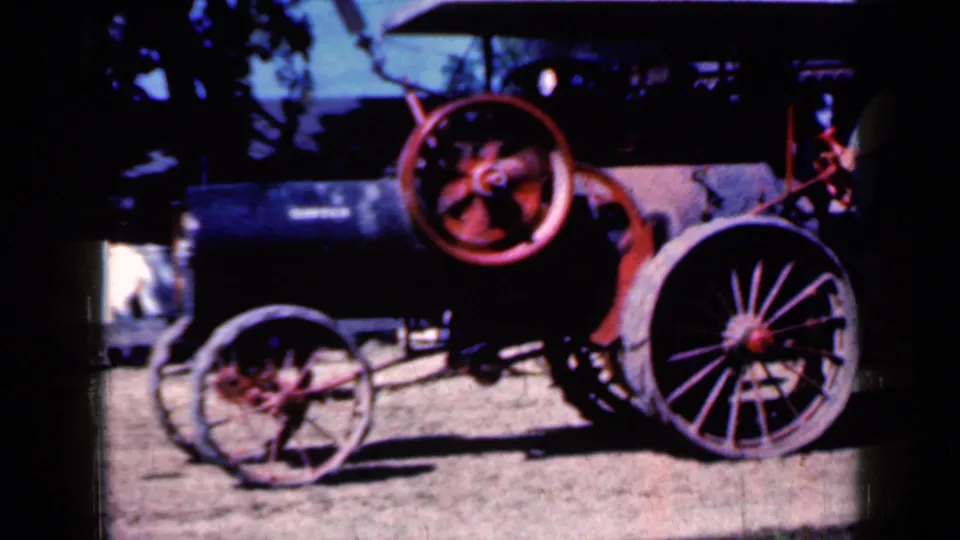 1959 Iowa home-movie frame of a weathered steam tractor, with the red flywheel prominent and a person partly visible near the machinery.