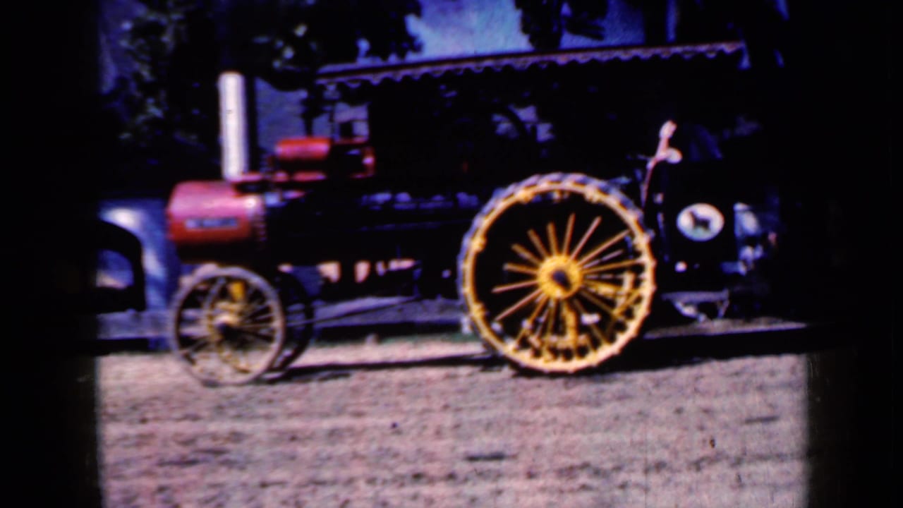 1959 Iowa frame of the steam tractor from the side, with the red boiler, pale smokestack, and large yellow spoked wheel visible against a shaded outdoor background.