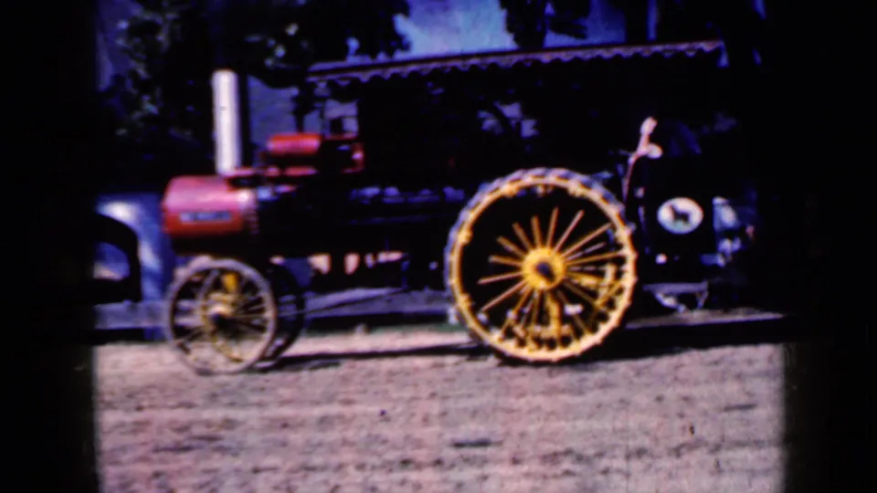 1959 Iowa frame of the steam tractor from the side, with the red boiler, pale smokestack, and large yellow spoked wheel visible against a shaded outdoor background.