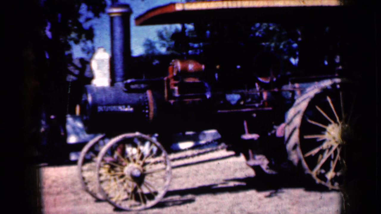 1959 Iowa frame showing the steam tractor at closer range, with the front wheel, smokestack, red boiler area, partial rear wheel, and a shaded structure overhead.