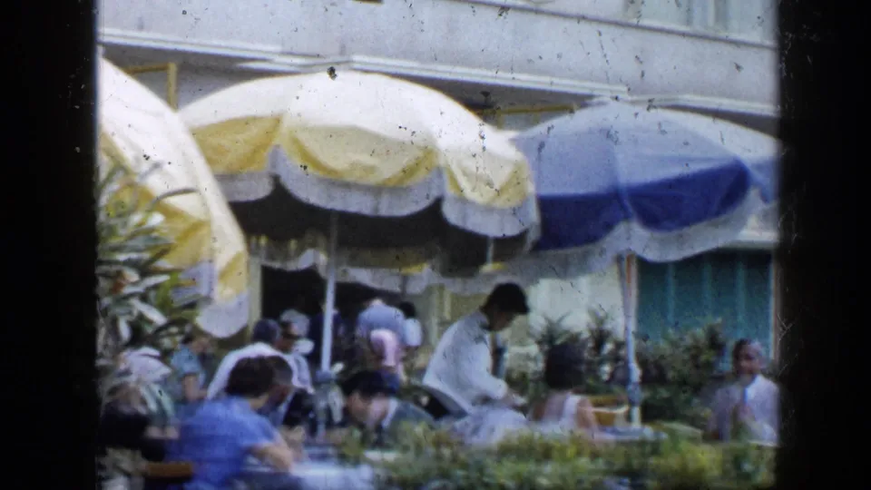 1959 Venezuela home-movie view of an outdoor restaurant terrace with diners, a waiter in white, umbrellas, and greenery.
