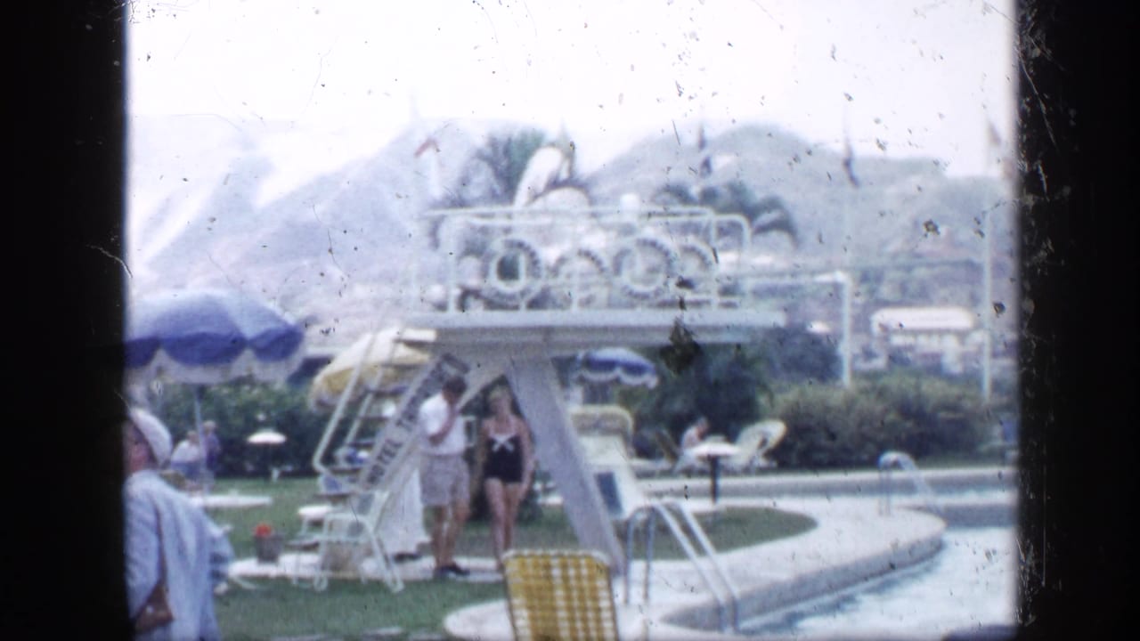 Poolside scene with a diving platform, two people standing nearby, umbrellas, and landscaped grounds.