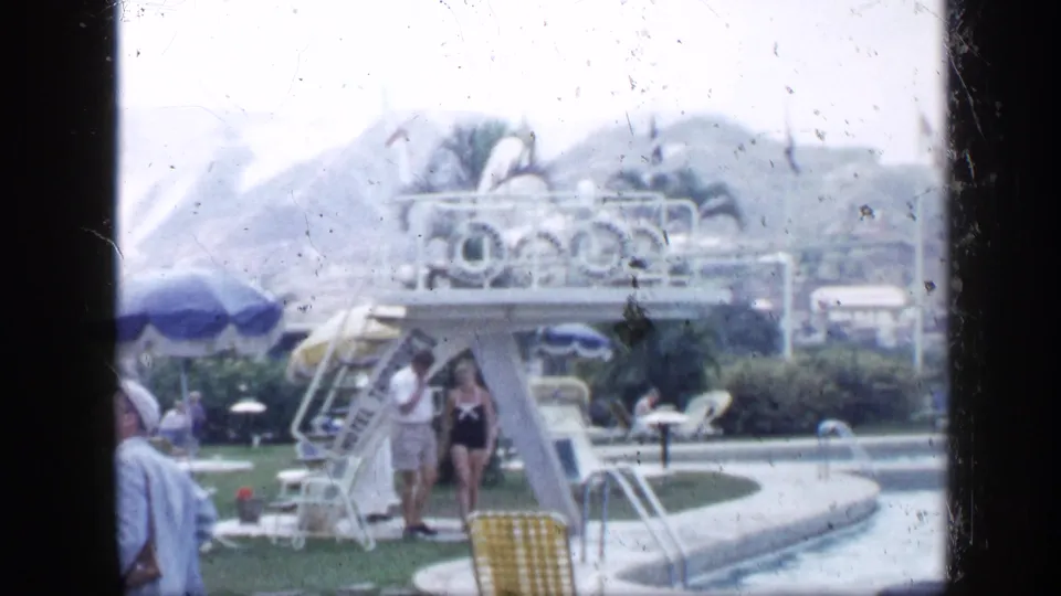1959 Venezuela resort pool area with guests near a diving board, patio umbrellas, palms, and curved pool edge.