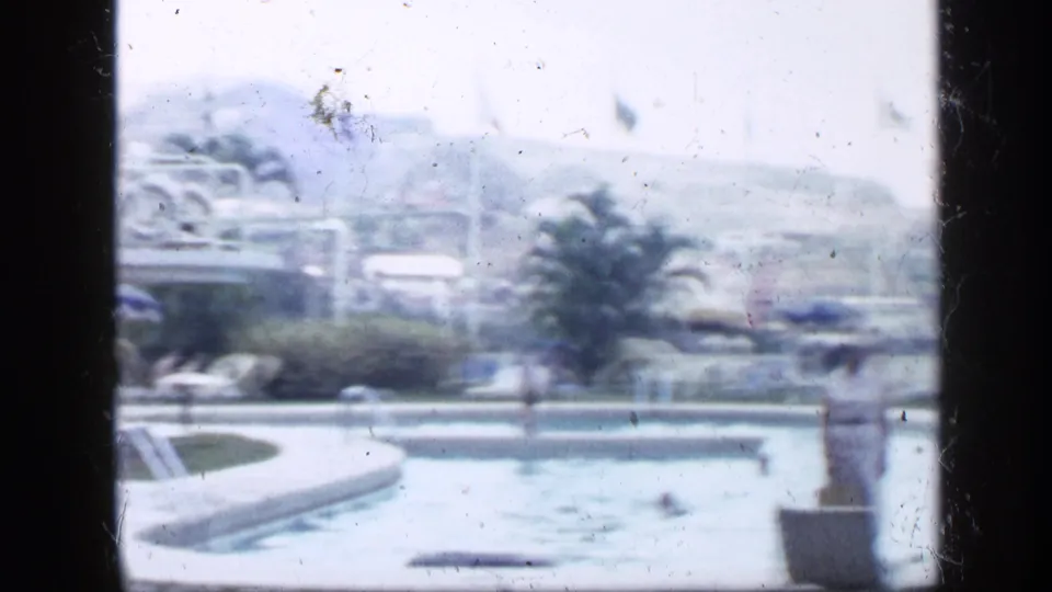 Soft 1959 Venezuela home-movie view across a resort pool toward a diving structure, palm, umbrellas, and surrounding grounds.