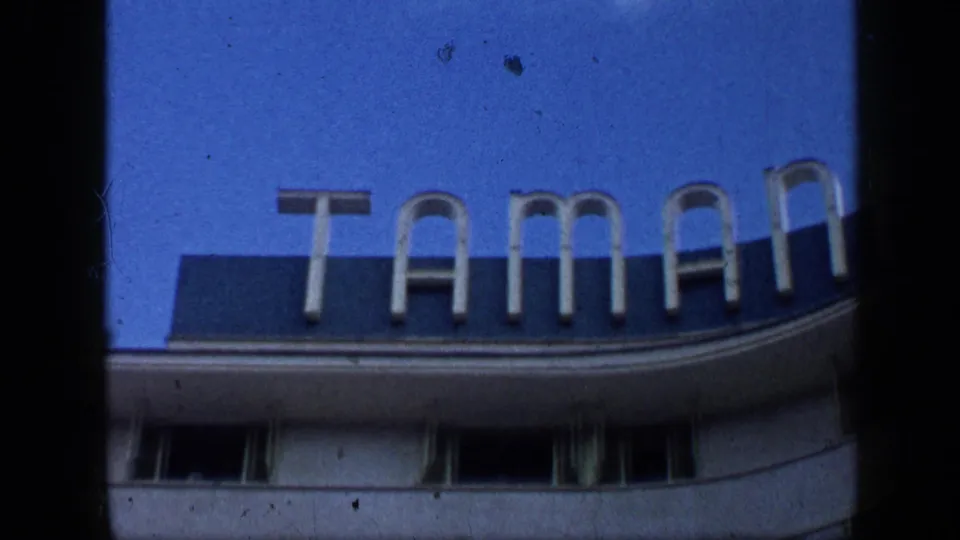1959 Venezuela exterior sign shot showing large TAMAN letters mounted above a curved building facade under blue sky.