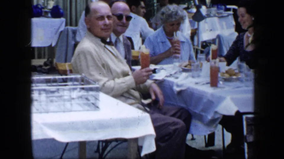 1959 Venezuela home-movie frame showing older adults dining outdoors, with a man in a light suit holding a drink.