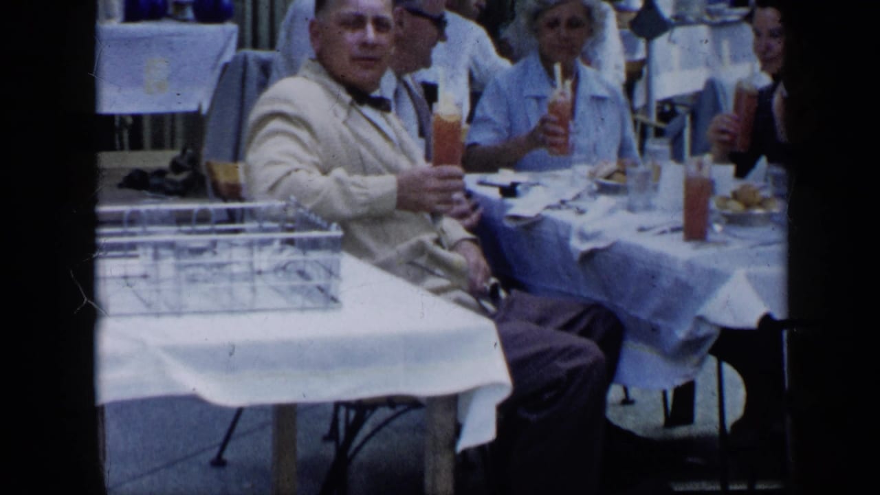 1959 Venezuela home-movie frame of adults at an outdoor table, with a man in a light suit holding a tall cocktail.