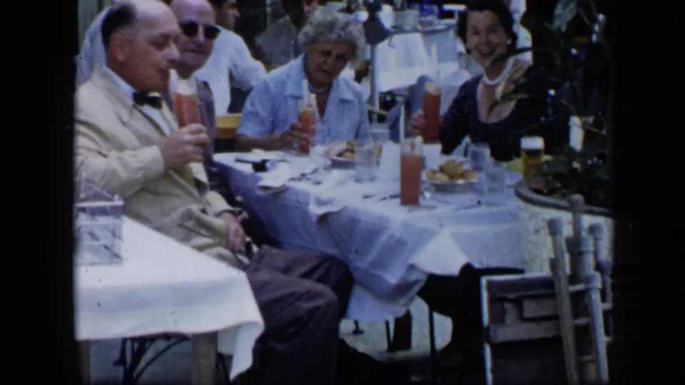 Adults at a 1959 Venezuela outdoor gathering hold cocktails and beer glasses while seated around a white table.