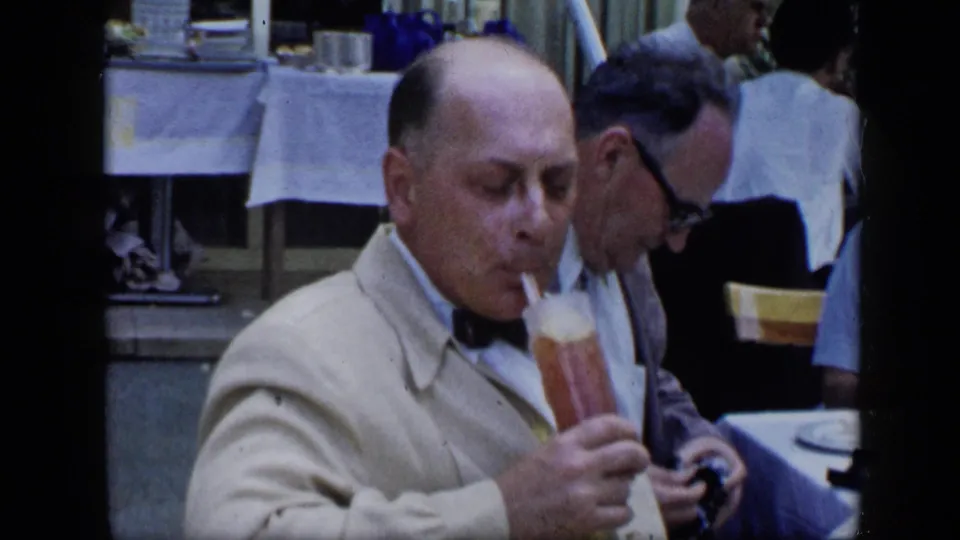 Close view of a man in a light suit and bow tie sipping a tall cocktail at the 1959 Venezuela outdoor gathering.