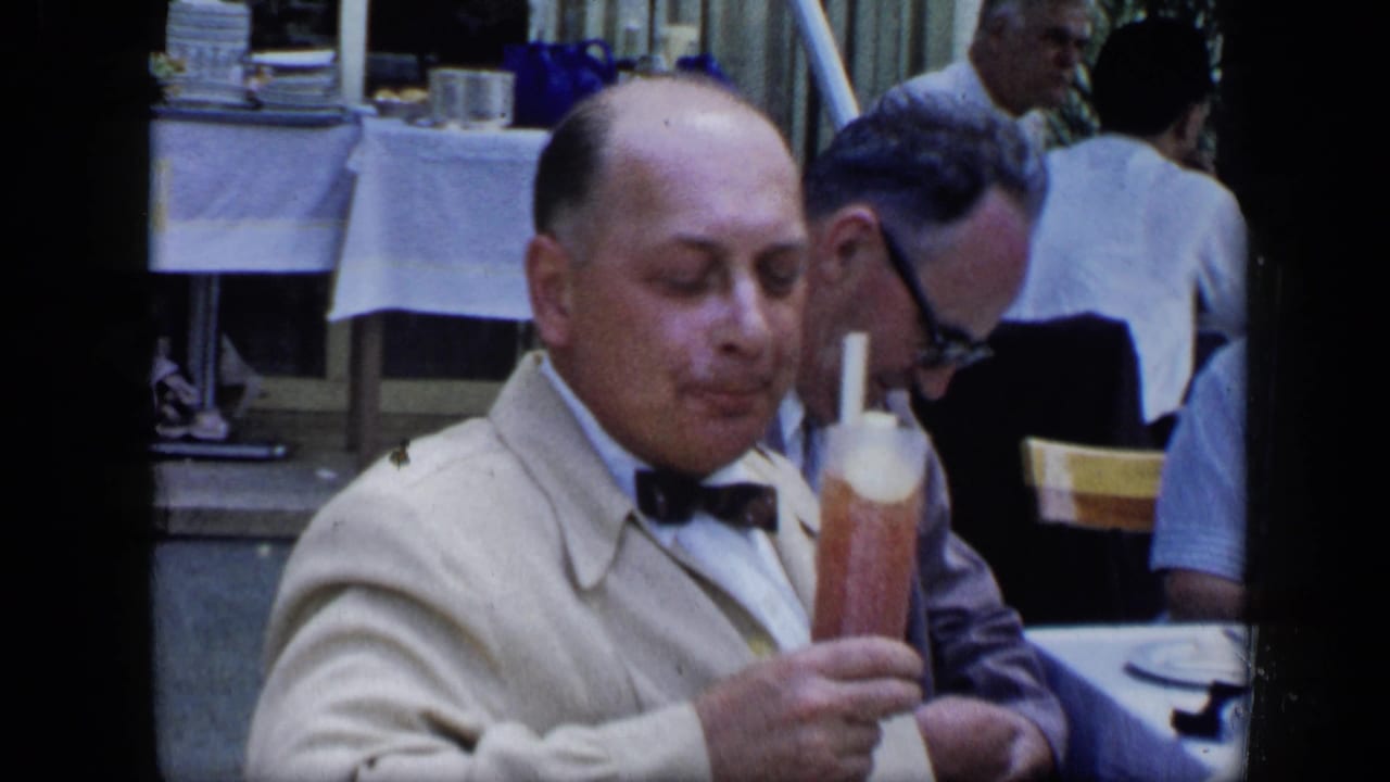 In 1959 Venezuela footage, a mature man in a light jacket and bow tie sits at a restaurant table with a tall drink and other diners behind him.