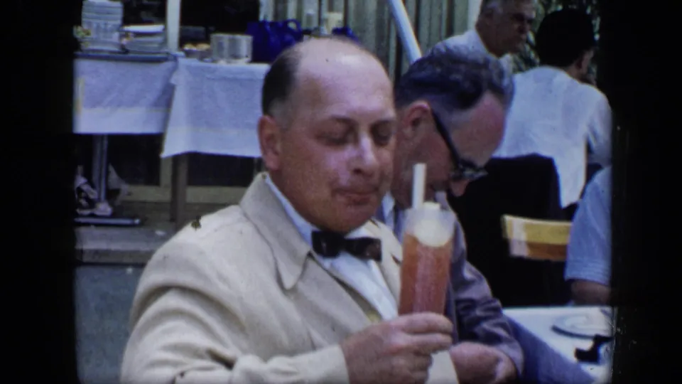 In 1959 Venezuela footage, a mature man in a light jacket and bow tie sits at a restaurant table with a tall drink and other diners behind him.