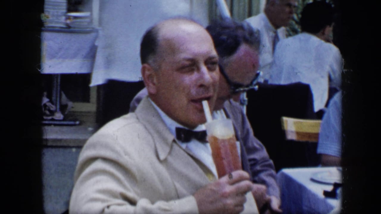 Man in a light jacket drinks through a straw at a restaurant table.