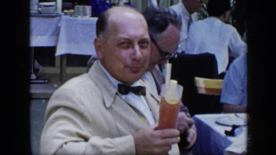 A mature man in a bow tie looks toward the camera while holding a tall drink at a restaurant table, with diners and table linens visible behind him.