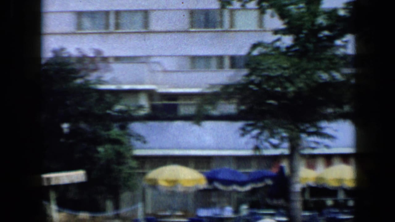 1959 Venezuela hotel exterior viewed from a shaded gallery, with trees and pool umbrellas near the lower edge.