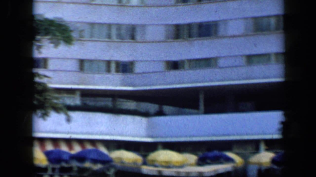 Curved hotel building with rows of windows above blue and yellow umbrellas by the pool area.