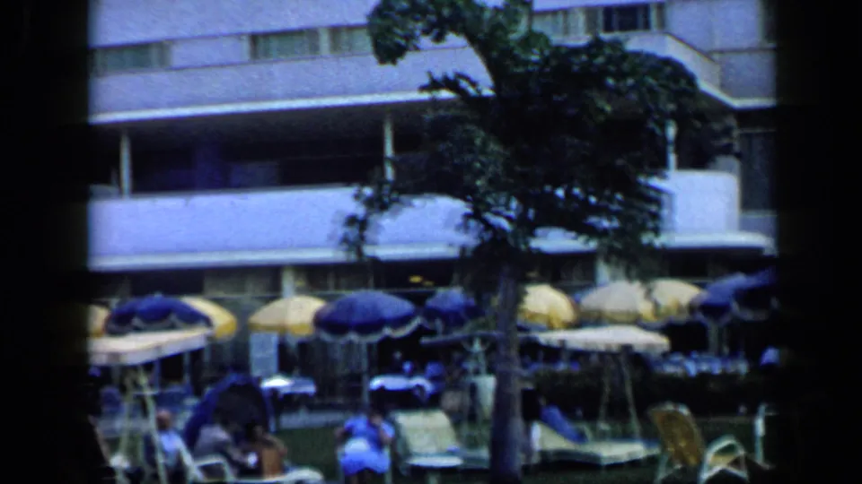 Poolside hotel area in 1959 Venezuela, with a palm tree, lounge chairs, umbrellas, and distant guests.