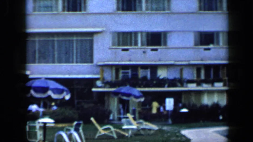 1959 Venezuela resort poolside view with lounge chairs, umbrellas, palm tree, and distant guests near a hotel facade.