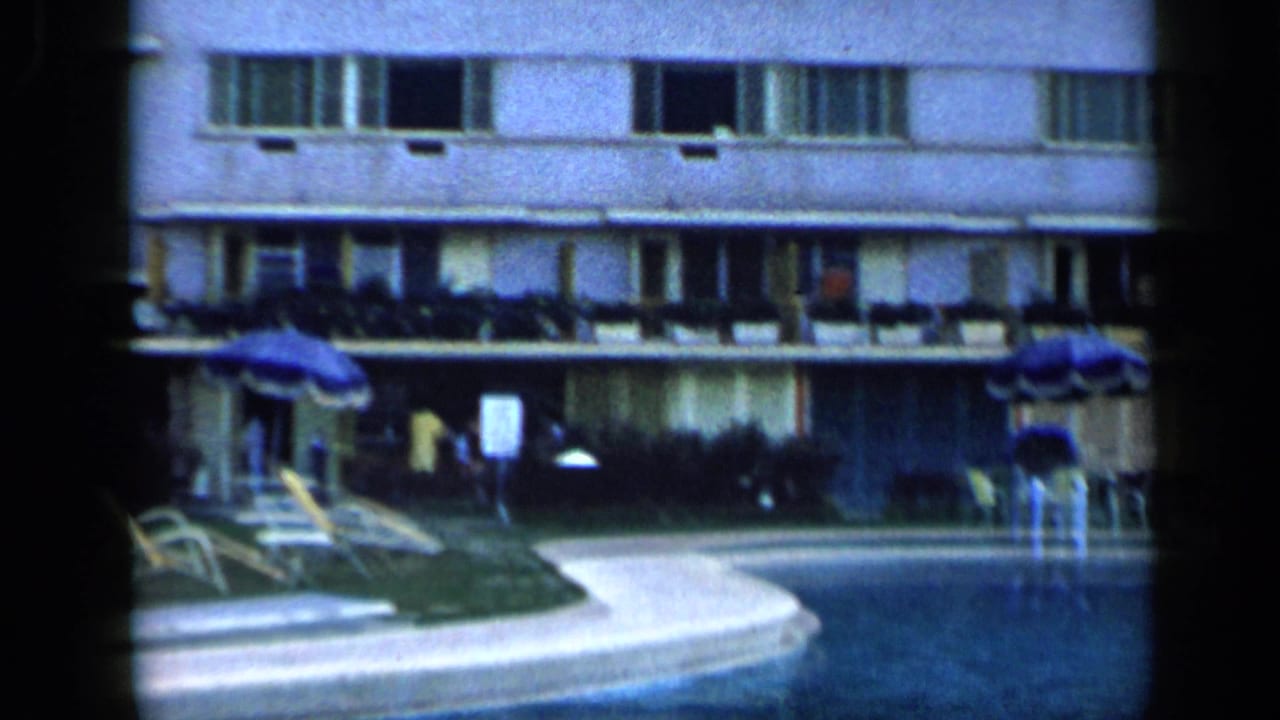 1959 Venezuela home-movie frame showing an outdoor hotel pool, lounge seating, umbrellas, balconies, and soft 8mm color grain.