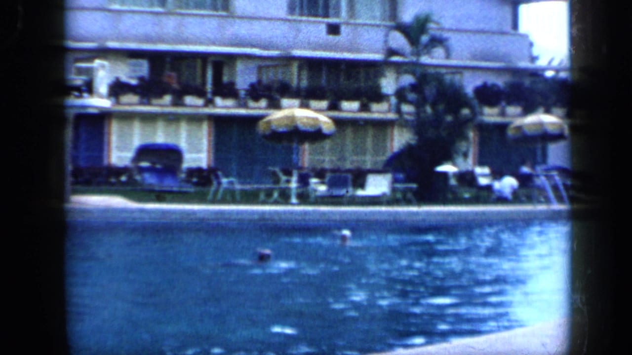 1959 Venezuela home-movie frame showing two distant swimmers in a hotel pool with umbrellas, lounge chairs, balconies, and soft color shift.