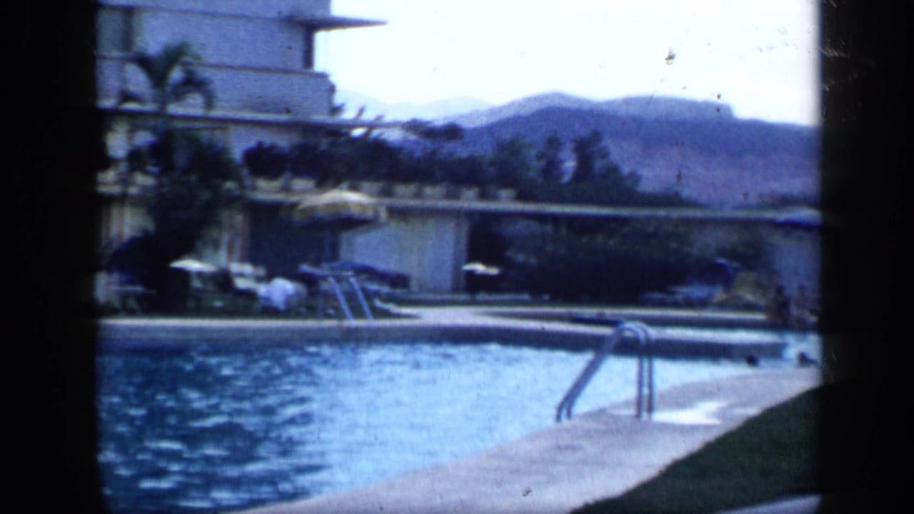 1959 Venezuela frame showing a hotel pool with distant swimmers, pool deck furniture, umbrellas, palm trees, and hills beyond the property.