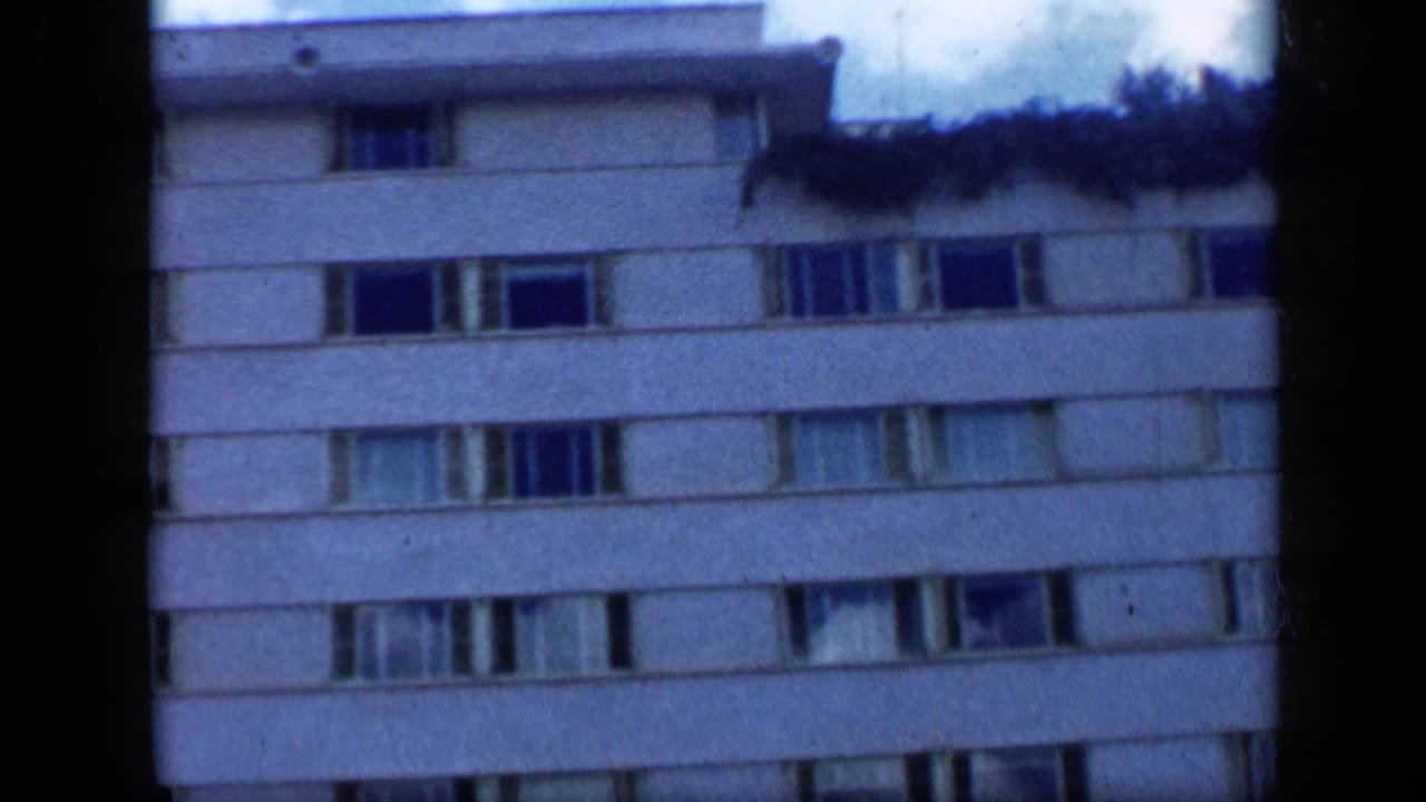 1959 Venezuela 8mm frame of the apartment building facade, showing multiple window rows, balcony edges, and rooftop vegetation.