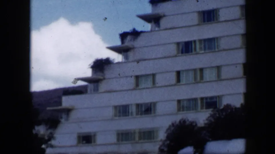 1959 Venezuela home-movie view of a white building seen from below, with balconies, window rows, clouds, and dark foliage at the base.